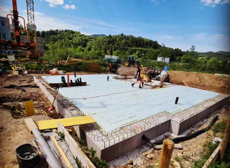 Baustelle mit Fundament, Arbeiter und Maschinen im Hintergrund. Vorbereitung für Bauprojekt in grüner Landschaft unter blauem Himmel.