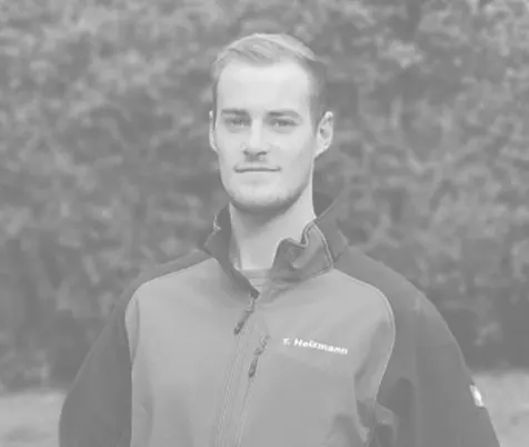 Tobias Heizmann in Arbeitsjacke im Freien, neutraler Hintergrund mit Sträuchern, entspannter Ausdruck.