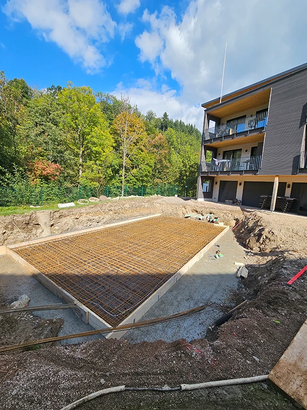 Baufortschritt: Betonfundament wird neben Wohngebäude für zukünftiges Projekt vorbereitet; grüne Waldkulisse im Hintergrund.