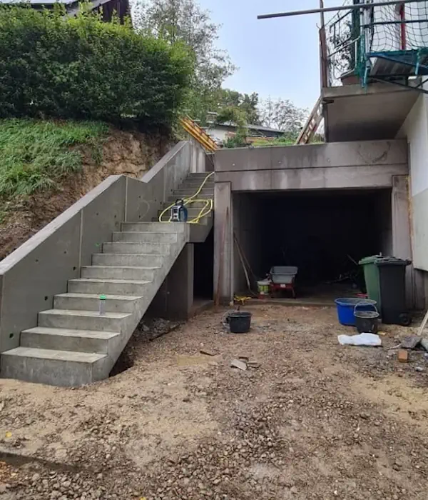 Baustelle mit Betonstufen und Einfahrt, Werkzeug und Baumaterialien verteilt. Gartengeländer und grüne Umgebung sichtbar.