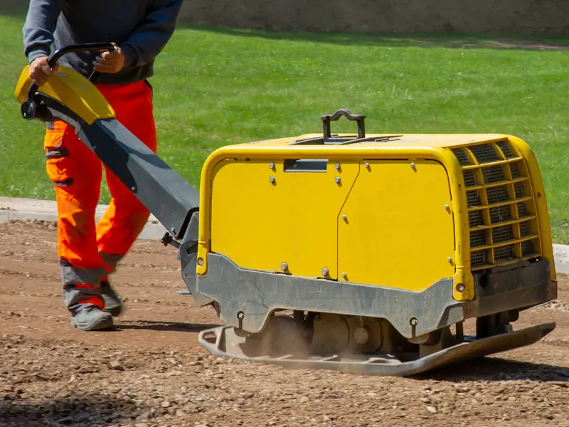 Mann bedient gelbe Bodenverdichtungsplatte auf Baustelle, angrenzend grüner Rasen. Effiziente Verdichtung für stabile Oberflächen.