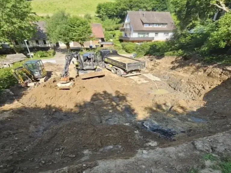 Bauarbeiten: Bagger und LKW graben Erdloch in Wohngebiet, umgeben von Bäumen und Häusern. Standort für neues Bauprojekt.