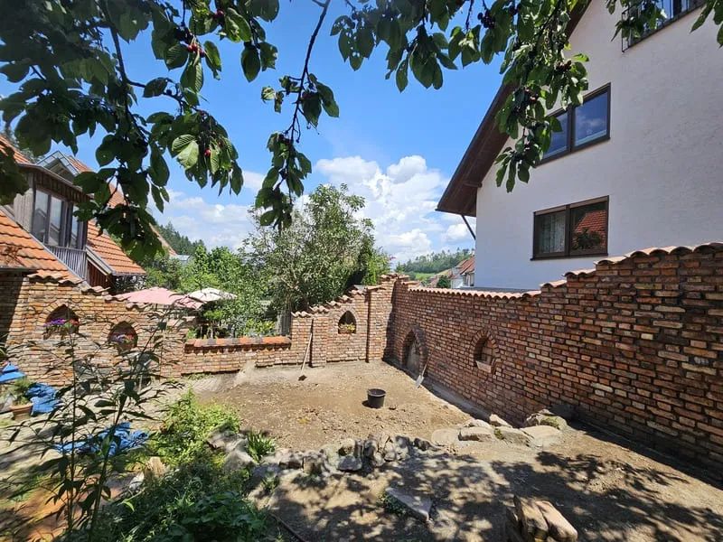 Gepflegter Garten mit Ziegelmauer, Bäumen und Haus im Hintergrund unter blauem Himmel, ideal für sommerliche Ruheplätze.