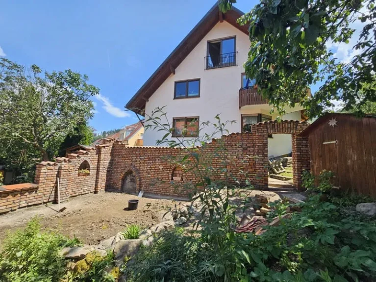 Modernes Einfamilienhaus mit Ziegelmauer im Garten, umgeben von Bäumen, sonniger Tag. Ideal für Familienleben und Ruhe im Grünen.