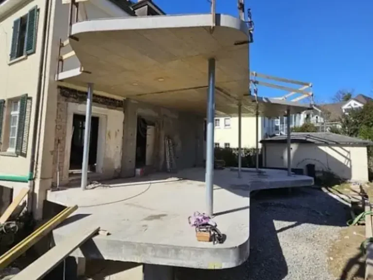 Hausanbau-Baustelle mit moderner Terrassen-Architektur und sichtbaren Stahlträgern, teilvergessene Arbeiten bei strahlendem Wetter.