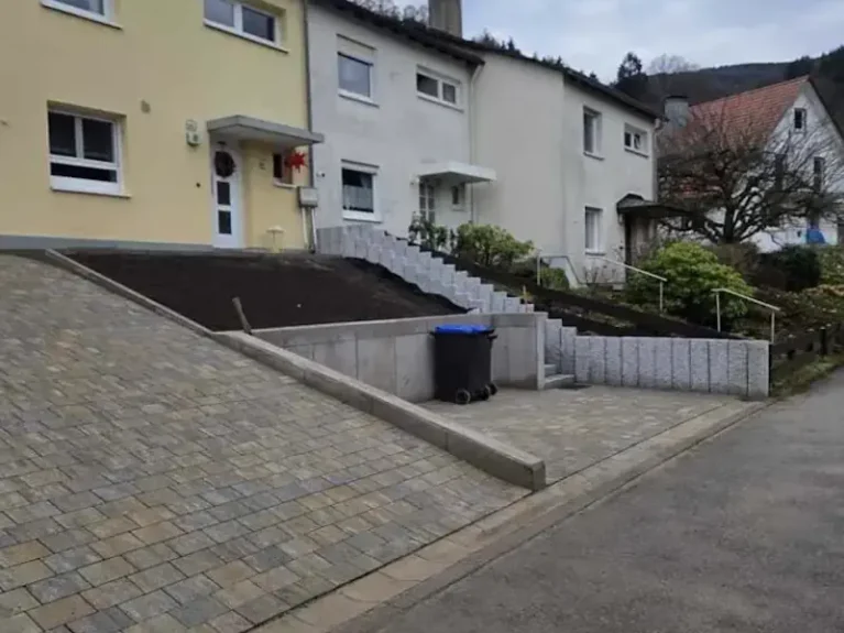 Reihenhaus mit modernem Pflasterweg und schräger Auffahrt, Treppe führt zum Eingangsbereich. Garten mit Pflanzen und Mülleimer rechts.