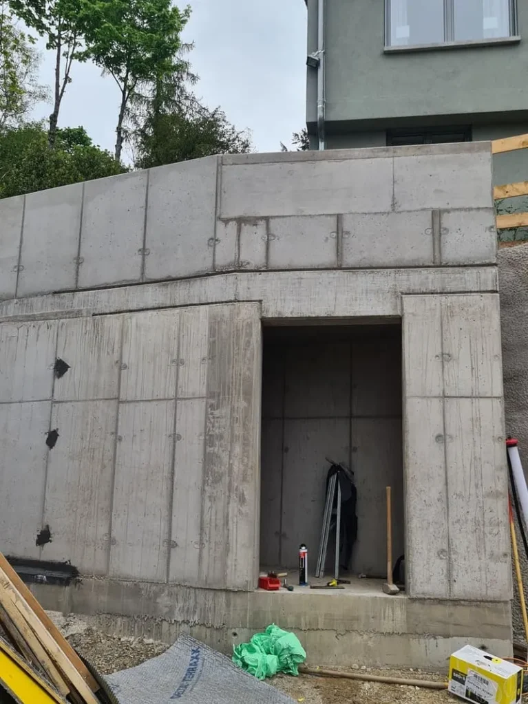 Betonwand mit rechteckiger Öffnung, Bauwerkzeug sichtbar, Wohnung im Hintergrund, Baustellenumgebung.