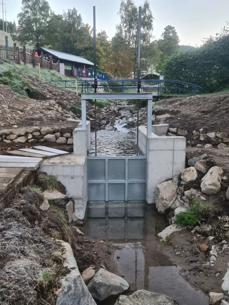 Bachlauf mit kleinem Wehr, umgeben von Natur, Steinen und einem Gebäude im Hintergrund. Ideal für Wasserregulierung oder landschaftliche Gestaltung.